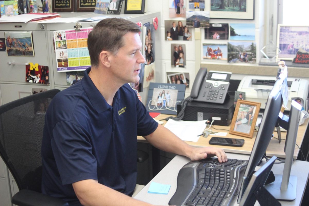 Middle school swim coach Aaron Portenga sits at his desk wearing a shirt embroidered with the words “Grand Haven Swimming” as he types on his computer. He oversees the middle school swim team sock sale fundraiser and helps bring more school spirit to the high school.